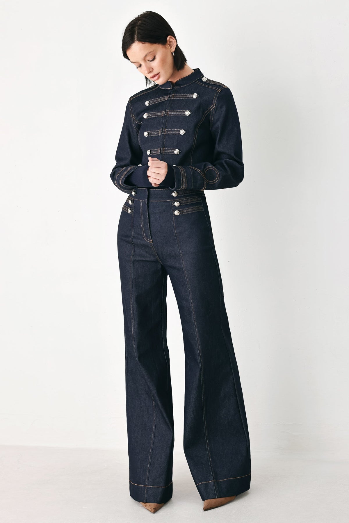 A model wearing a dark navy blue denim jumpsuit with military-style double-breasted buttons, photographed against a clean white studio background.