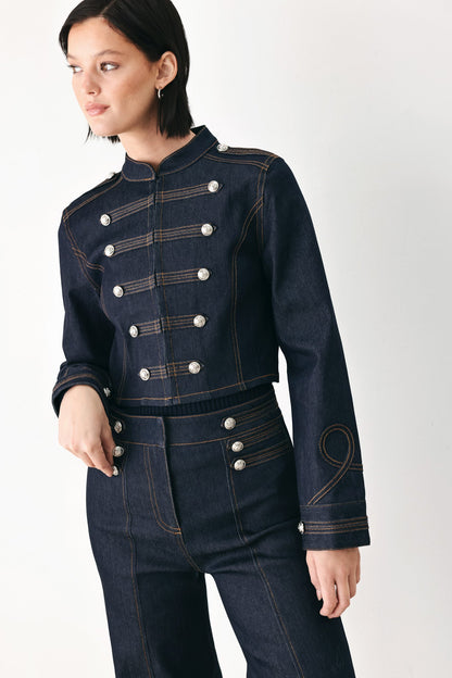 A model wearing a dark blue denim military-style jacket and matching high-waisted jeans, featuring double-breasted silver buttons and contrast stitching, set against a plain light gray background.