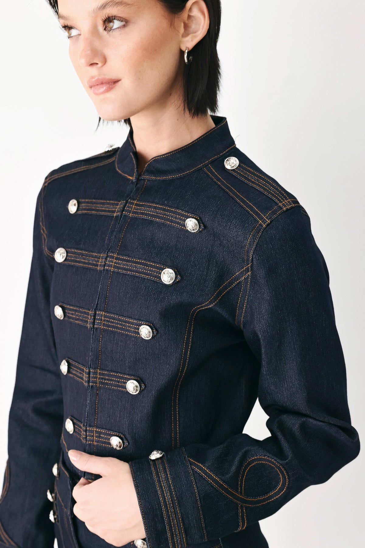A close-up of a model wearing a dark navy blue denim jacket with double-breasted silver buttons and contrasting gold-colored stitching, set against a plain light gray background.