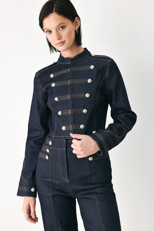 A model wearing a dark indigo denim military-style jacket with double-breasted silver buttons and matching dark denim trousers, photographed against a clean white studio background.