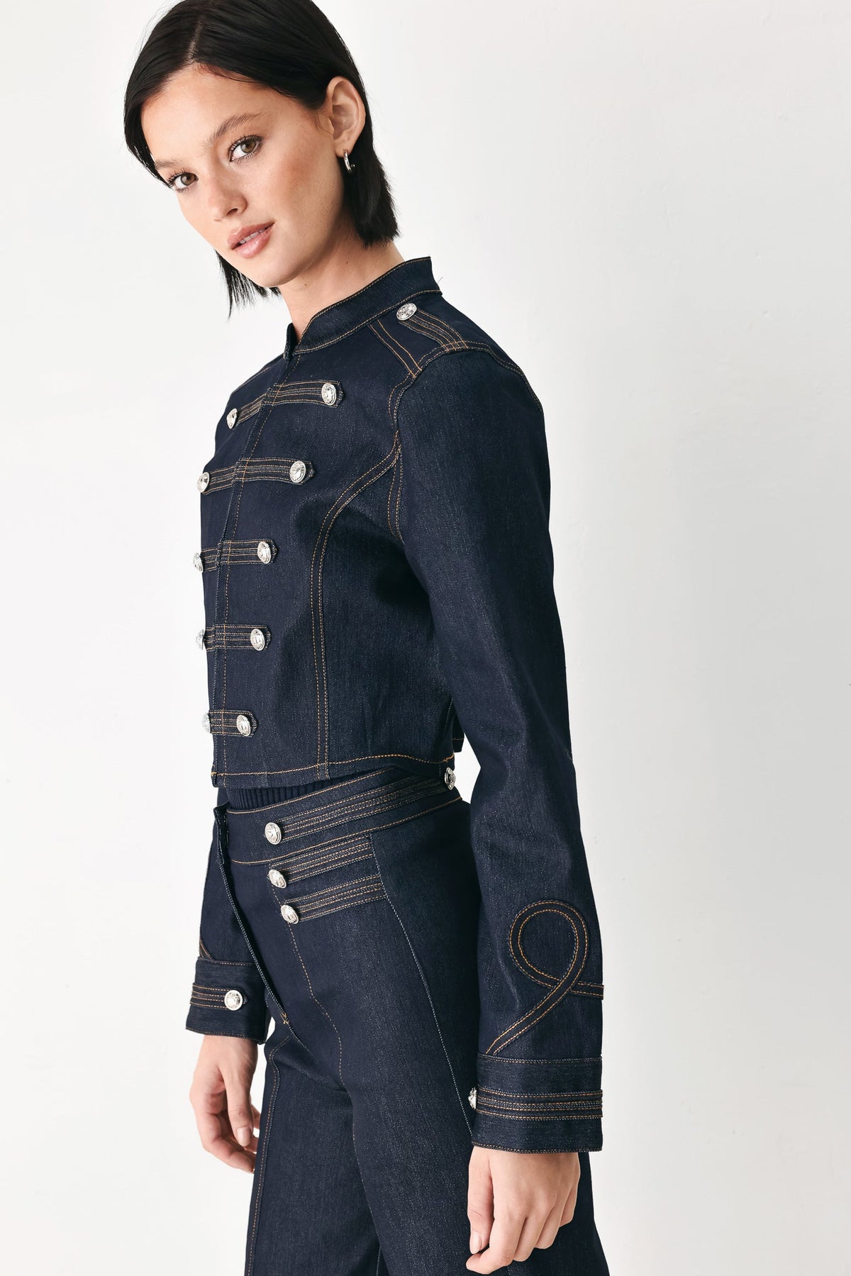 A model wearing a dark navy blue denim military-style jacket with silver buttons and contrast stitching, paired with matching dark denim trousers, against a plain light gray studio background.