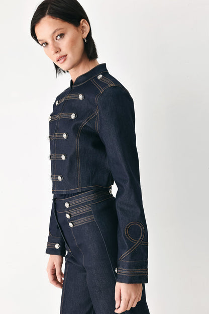 A model wearing a dark navy blue denim military-style jacket with silver buttons and contrast stitching, paired with matching dark denim trousers, against a plain light gray studio background.