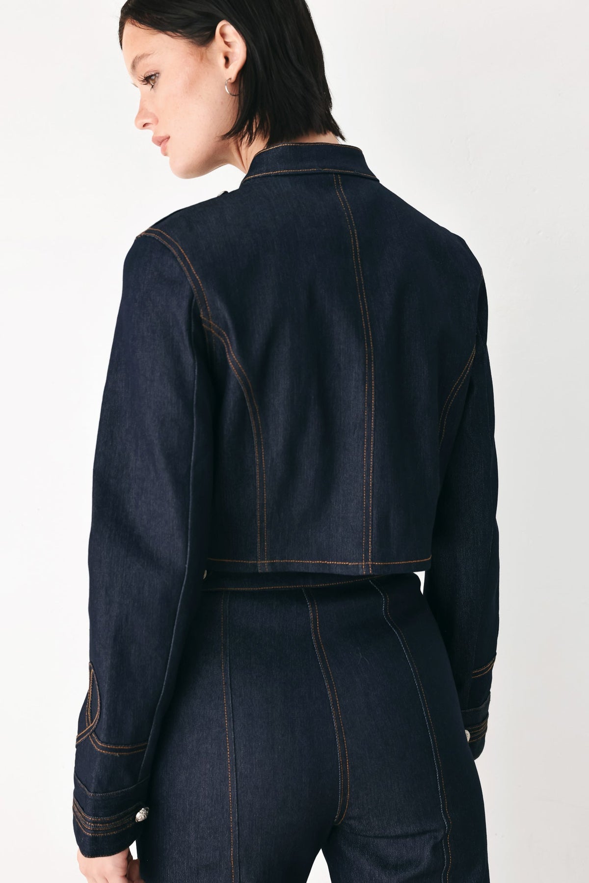 A model shown from the back wearing a dark indigo denim jacket and matching high-waisted denim pants, featuring contrast orange-brown stitching, against a plain white studio background.