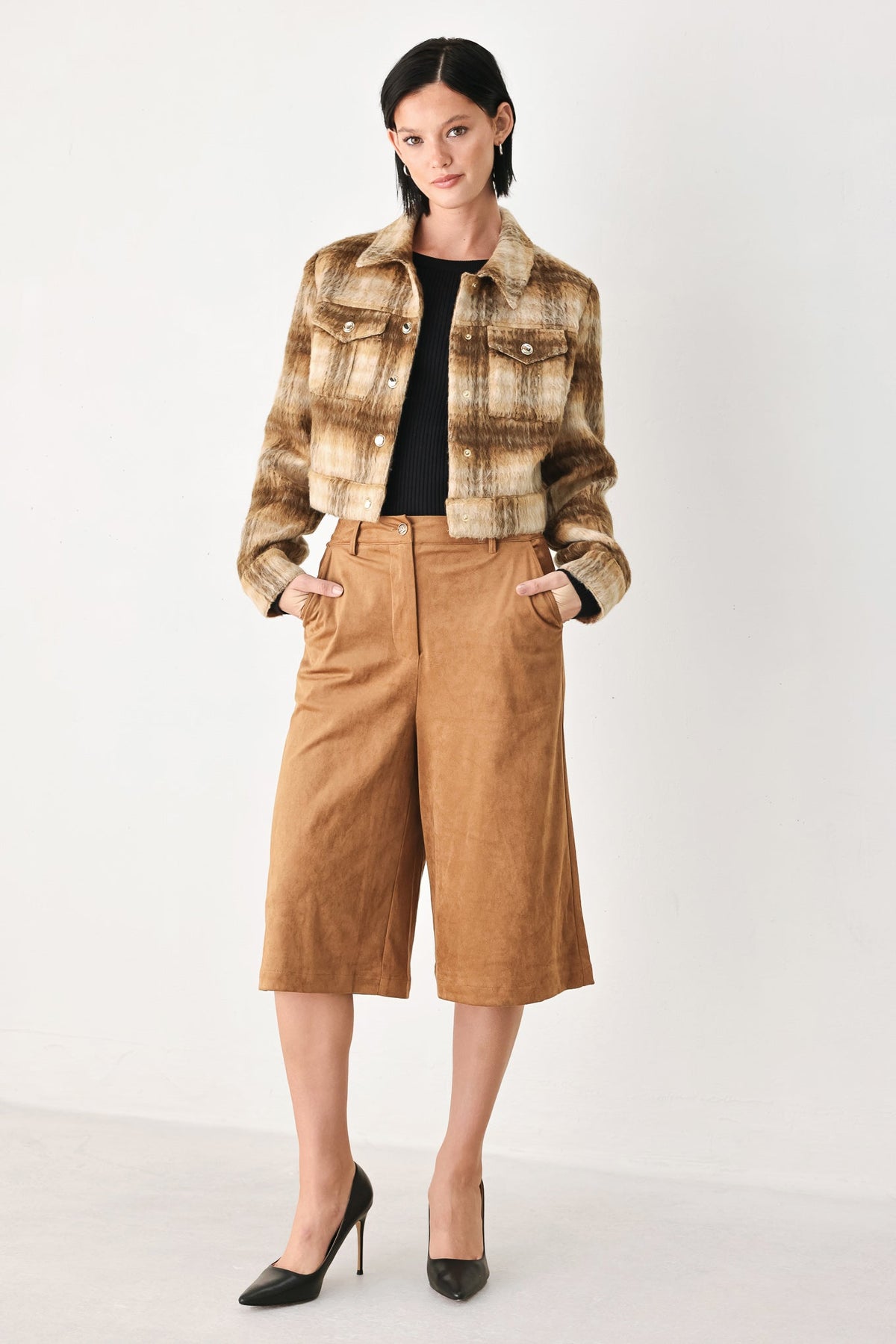 A model stands against a plain white studio background wearing a cropped brown and beige marled jacket over a black top, paired with tan wide-leg cropped pants and black pointed heels.