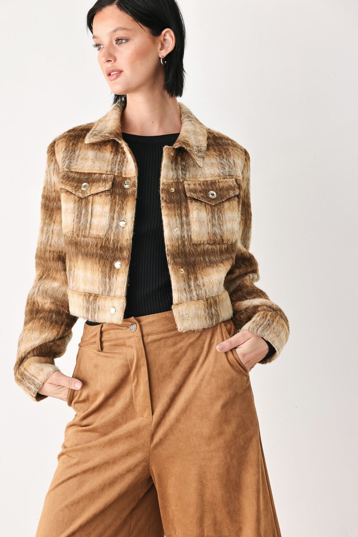 A woman models a cropped, long-sleeve plaid jacket in brown and beige tones, worn over a black top and paired with brown trousers, against a plain light background.