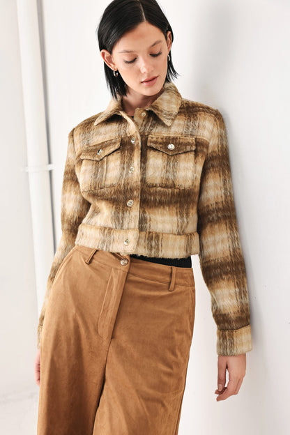 A woman wearing a cropped, long-sleeve button-up jacket with a brown and beige marled texture, paired with brown corduroy trousers, standing against a white wall.