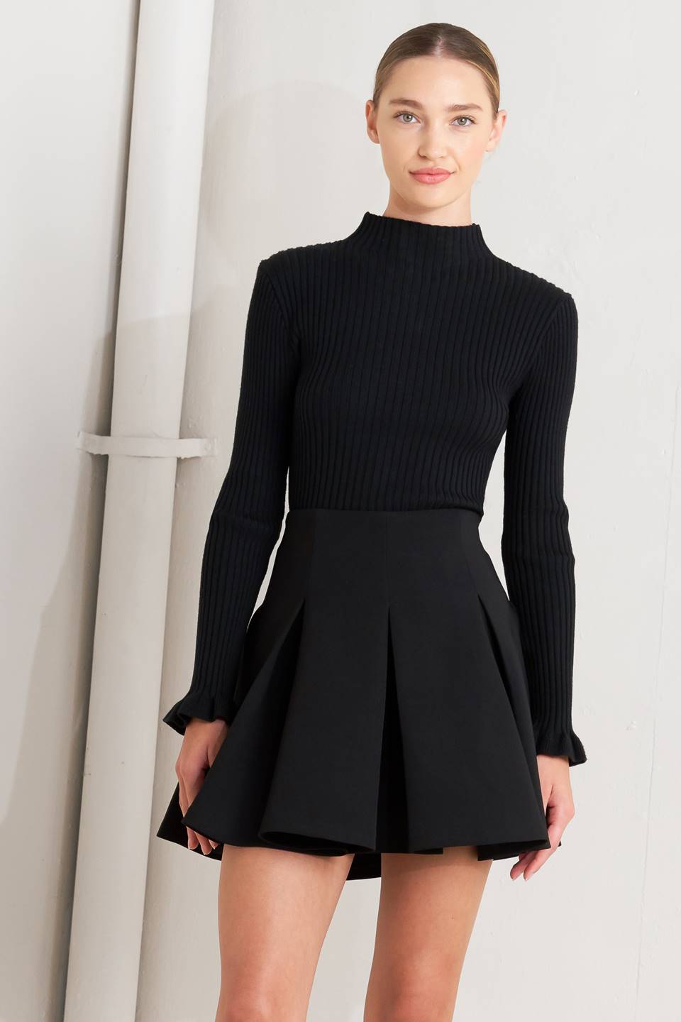 A woman modeling a black ribbed long-sleeve mock neck top paired with a matching black pleated mini skirt, standing against a light gray studio backdrop.