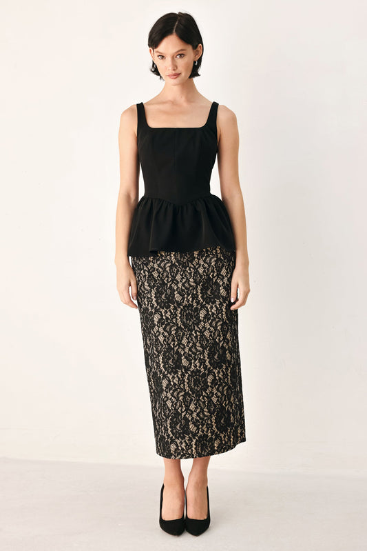 A model wearing a black sleeveless peplum top and a black lace midi skirt, paired with black heels, standing against a plain off-white studio background.
