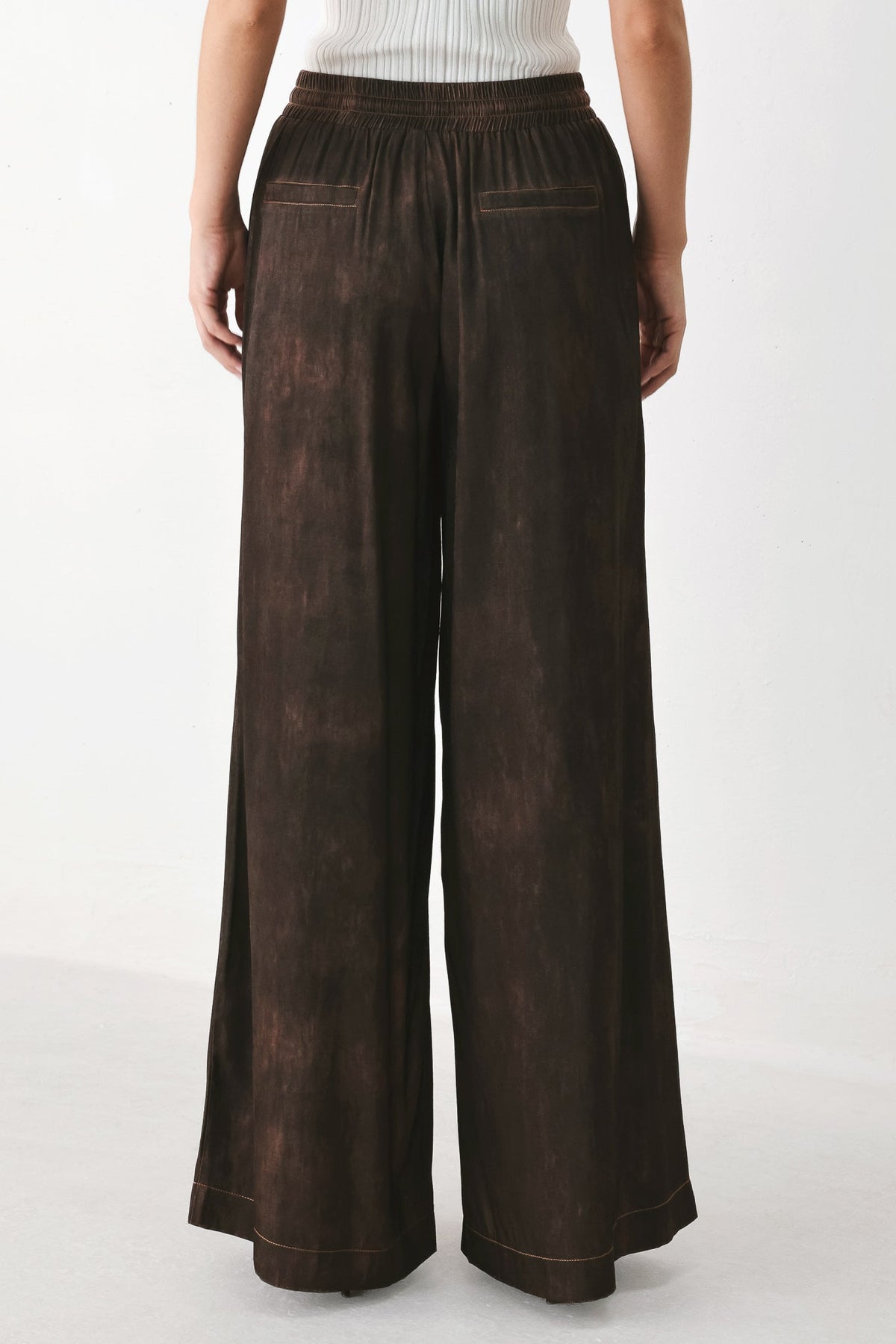 A person shown from the back wearing dark brown wide-leg pants with a mottled, washed texture and an elastic waistband, paired with a white ribbed top against a plain light background.