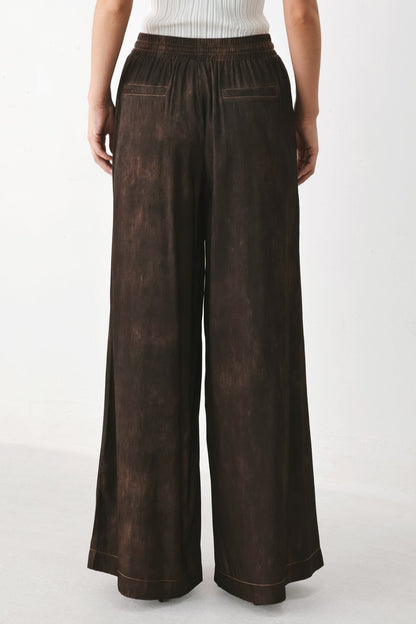 A person shown from the back wearing dark brown wide-leg pants with a mottled, washed texture and an elastic waistband, paired with a white ribbed top against a plain light background.