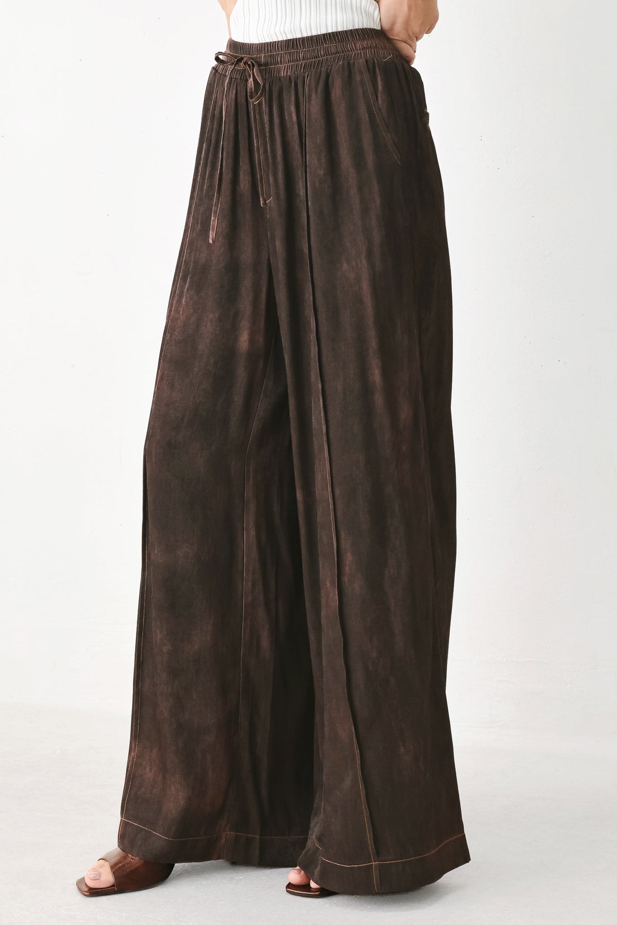 A person wearing loose, wide-leg brown pants with a drawstring waist and cuffed hems, shown from the waist down against a plain light background.