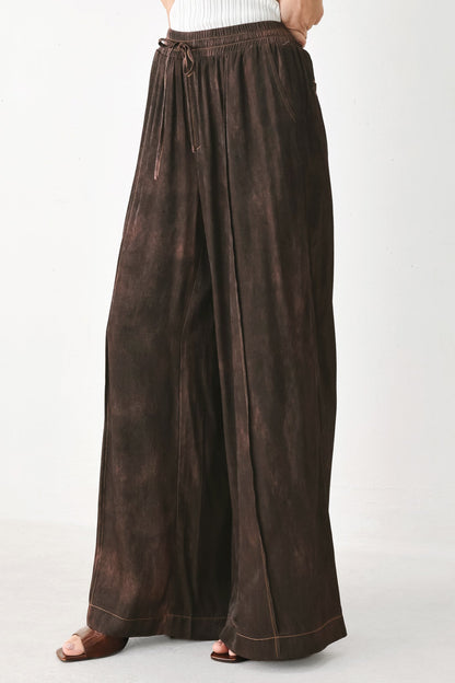 A person wearing loose, wide-leg brown pants with a drawstring waist and cuffed hems, shown from the waist down against a plain light background.