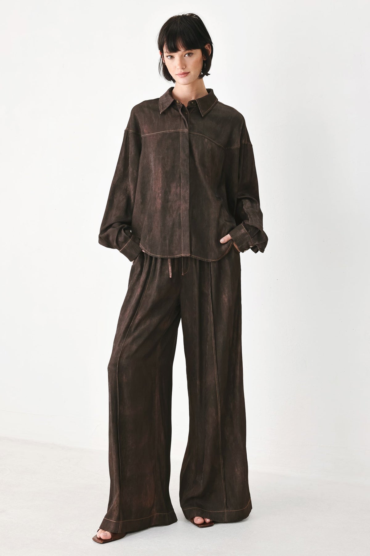 A model wearing a dark brown matching lounge set consisting of a long-sleeve button-up shirt and wide-leg trousers, photographed against a clean white studio background.