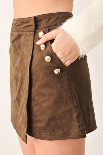 A close-up of a brown suede mini skirt with gold-tone decorative buttons down the front side, worn by a model whose hand is tucked into the pocket.