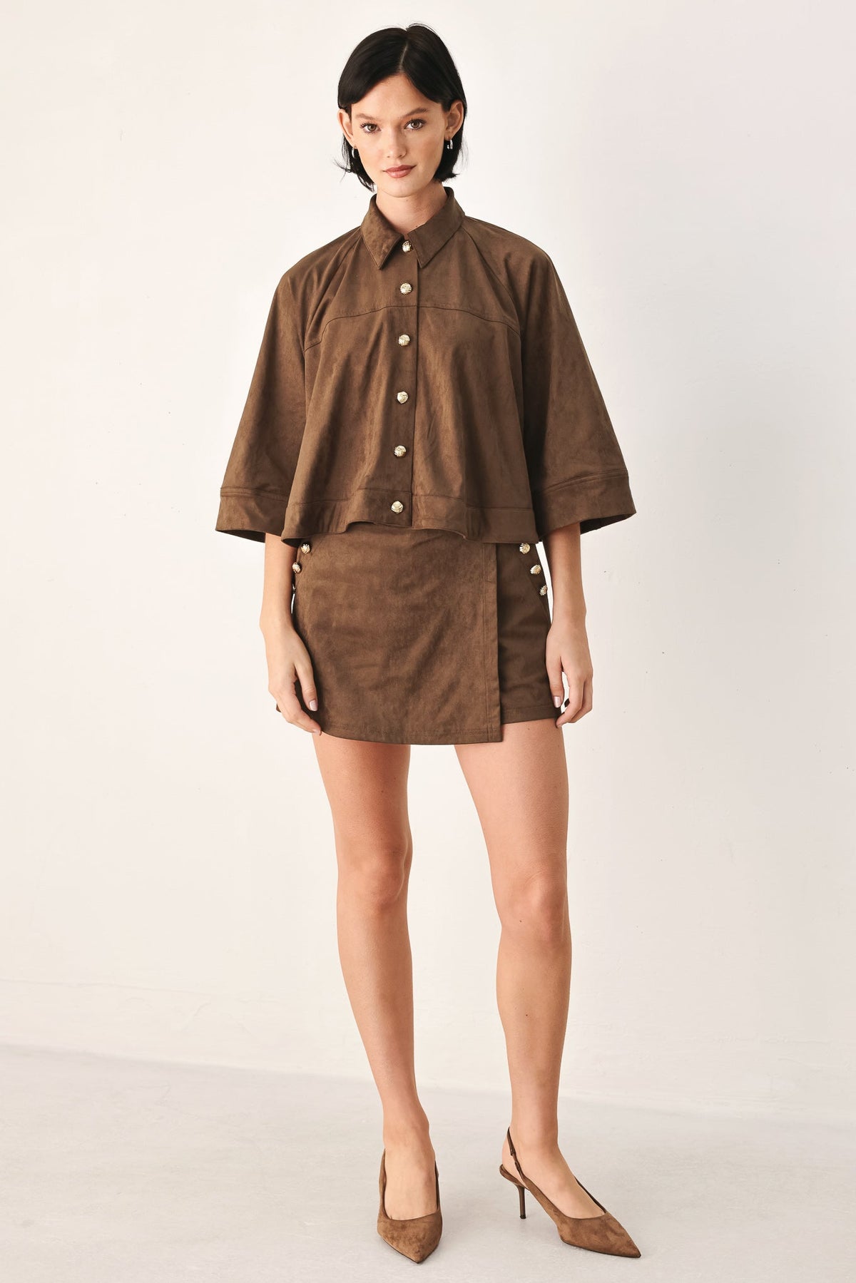 A model wearing a matching brown cropped button-front shirt and mini skirt set with side buttons, paired with brown pointed-toe heels, against a plain off-white studio background.