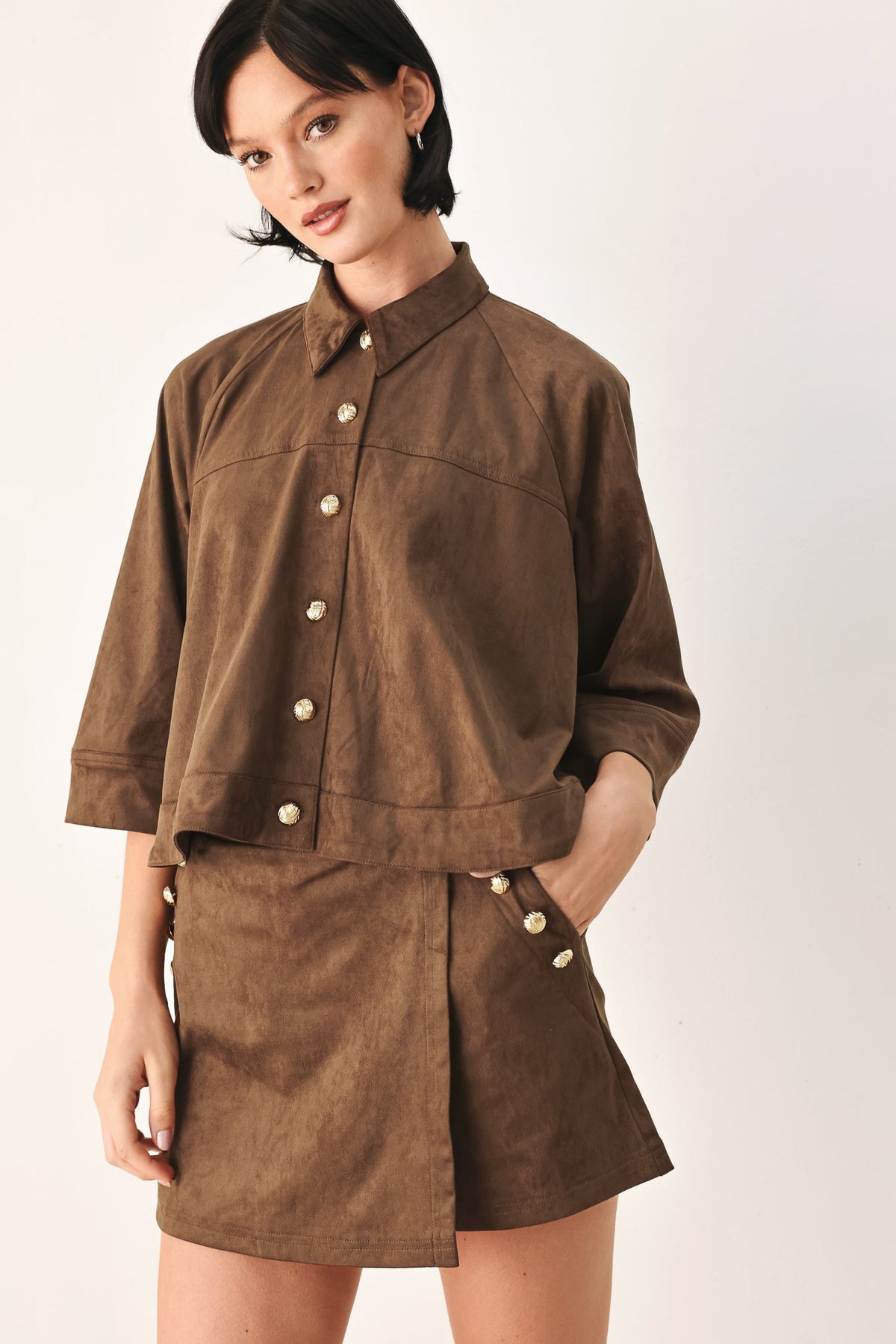 A model wearing a brown suede-like button-front shirt jacket and matching shorts with light-colored buttons, photographed against a plain light background.