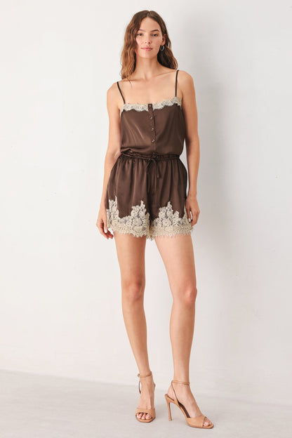 A woman modeling a brown satin romper with white lace trim at the neckline and hem, paired with nude high-heeled sandals against a plain white studio background.