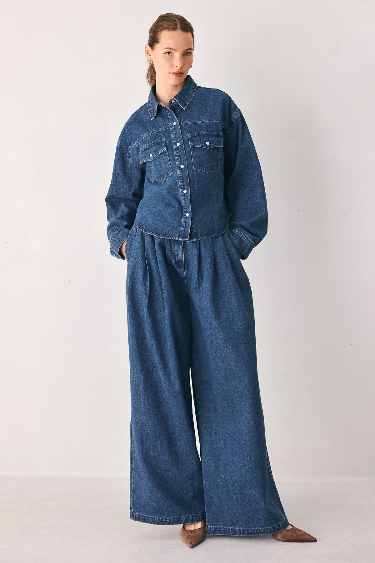 High-rise medium indigo wide-leg denim pants worn with a matching denim shirt jacket in a light gray studio.