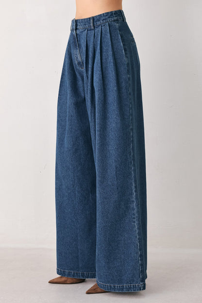 Model wearing high-rise pleated denim pants in medium indigo against a plain light gray background.