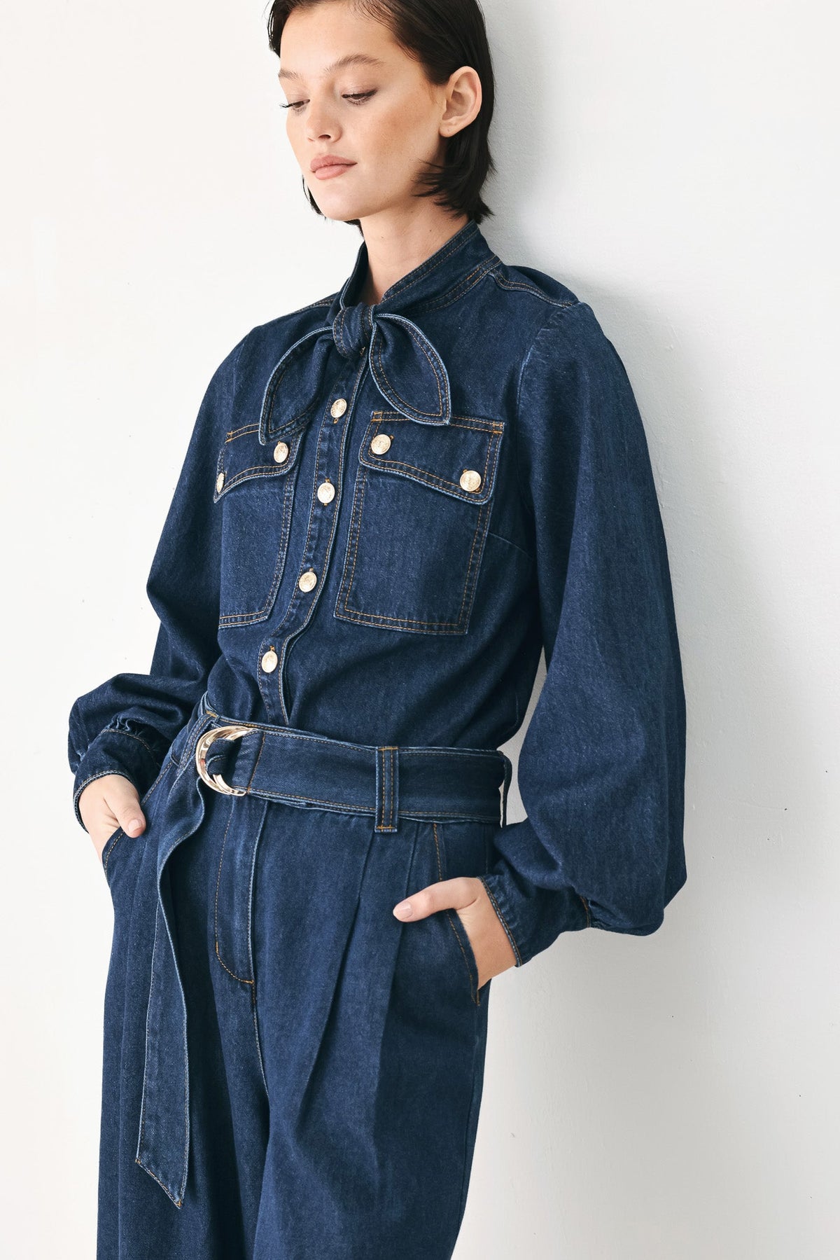 A model wearing a dark blue denim jumpsuit with a tie-neck detail, silver snap buttons, and a matching denim belt with a silver buckle, standing against a plain off-white wall.