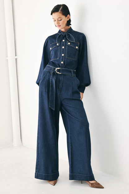A model wearing a dark blue denim jumpsuit with a tie-neck, button details, and a matching tie belt, paired with brown pointed-toe heels, standing against a white wall.