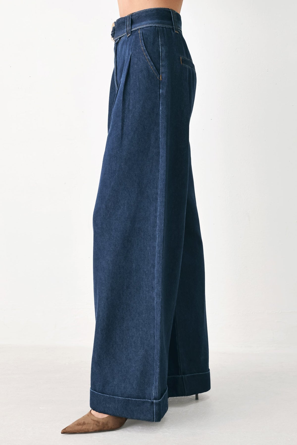 A person wearing dark blue wide-leg pleated denim jeans with cuffed hems, paired with brown pointed-toe heels, shown from the waist down against a plain light background.