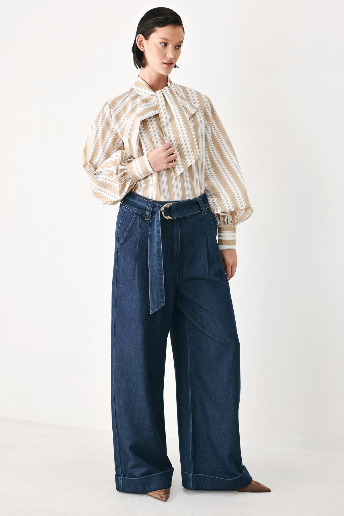 A model wearing a cream and beige vertically striped long-sleeve blouse with a large bow collar, tucked into dark blue wide-leg denim jeans with a matching belt.