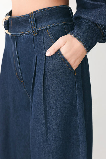 Close-up of a person wearing dark blue denim high-waisted wide-leg pants with pleats, a matching belt, and a long-sleeve denim top, with one hand in the pocket.