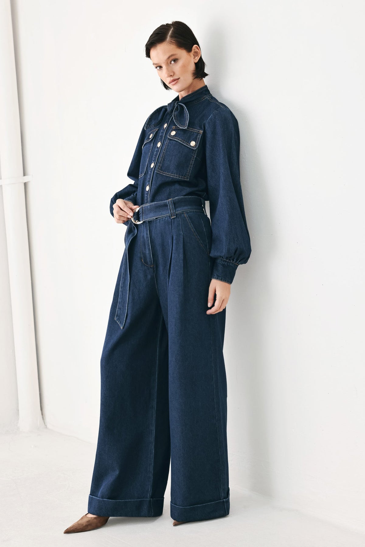 A model wearing a dark blue denim jumpsuit with a high collar, long sleeves, and wide legs, standing against a white studio wall.