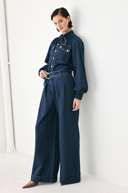 A model wearing a dark blue denim jumpsuit with a high collar, long sleeves, and wide legs, standing against a white studio wall.