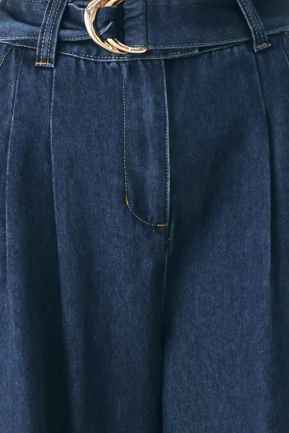 Close-up of dark blue denim trousers with pleats, contrast stitching, and a gold-tone belt buckle at the waist.
