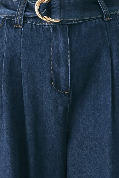 Close-up of dark blue denim trousers with pleats, contrast stitching, and a gold-tone belt buckle at the waist.