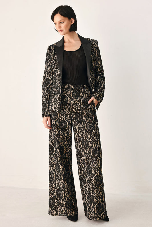 A model wearing a matching black and beige floral lace blazer and wide-leg trousers set, styled with a black ribbed top, against a plain off-white studio background.