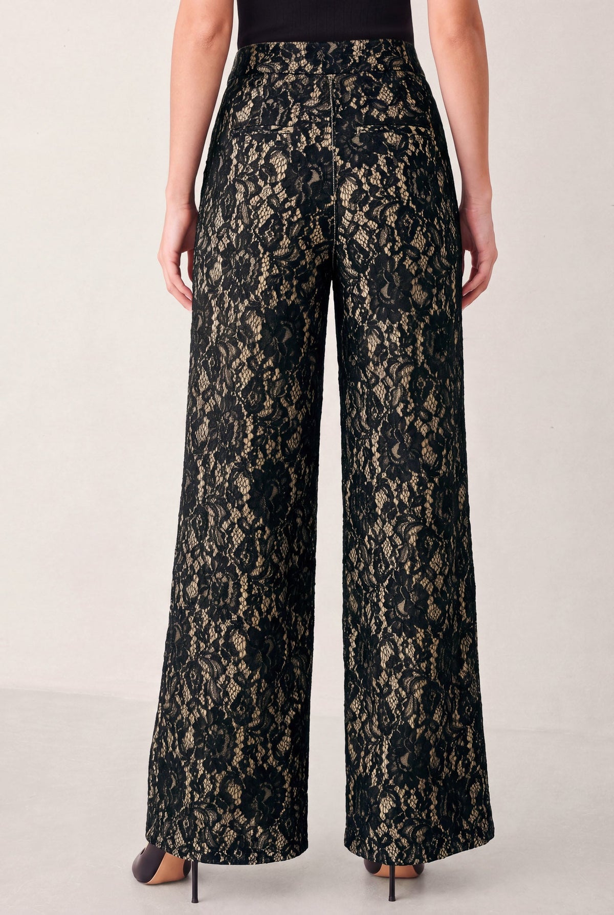 Black lace wide-leg pants with a sheer floral pattern, shown on a model from the back against a light gray studio background.