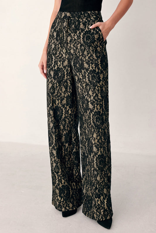 A model wearing high-waisted, wide-leg trousers featuring a black and beige floral lace pattern, shown from the waist down against a plain light background.