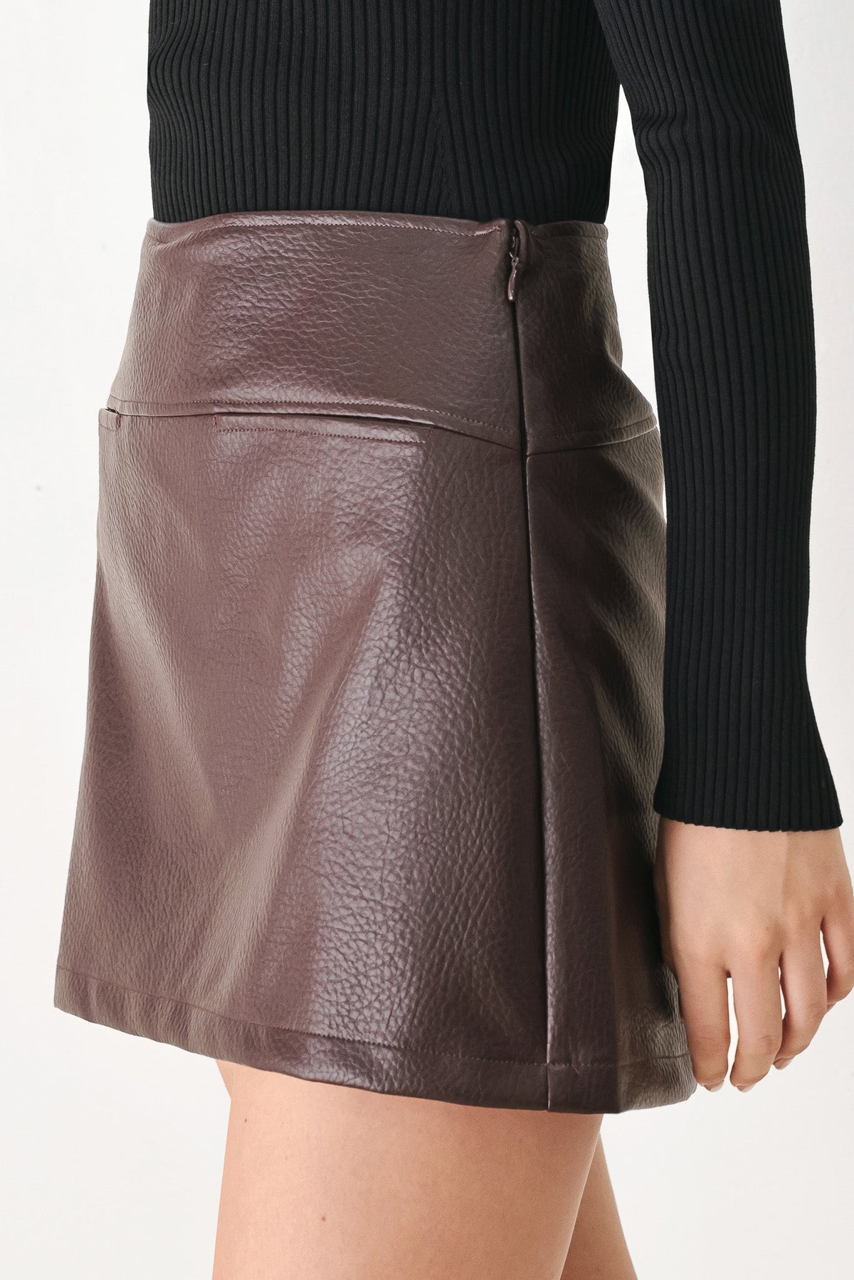 A close-up of a model wearing a brown faux-leather mini skirt with a pebbled texture, paired with a black ribbed long-sleeve top, against a plain white background.