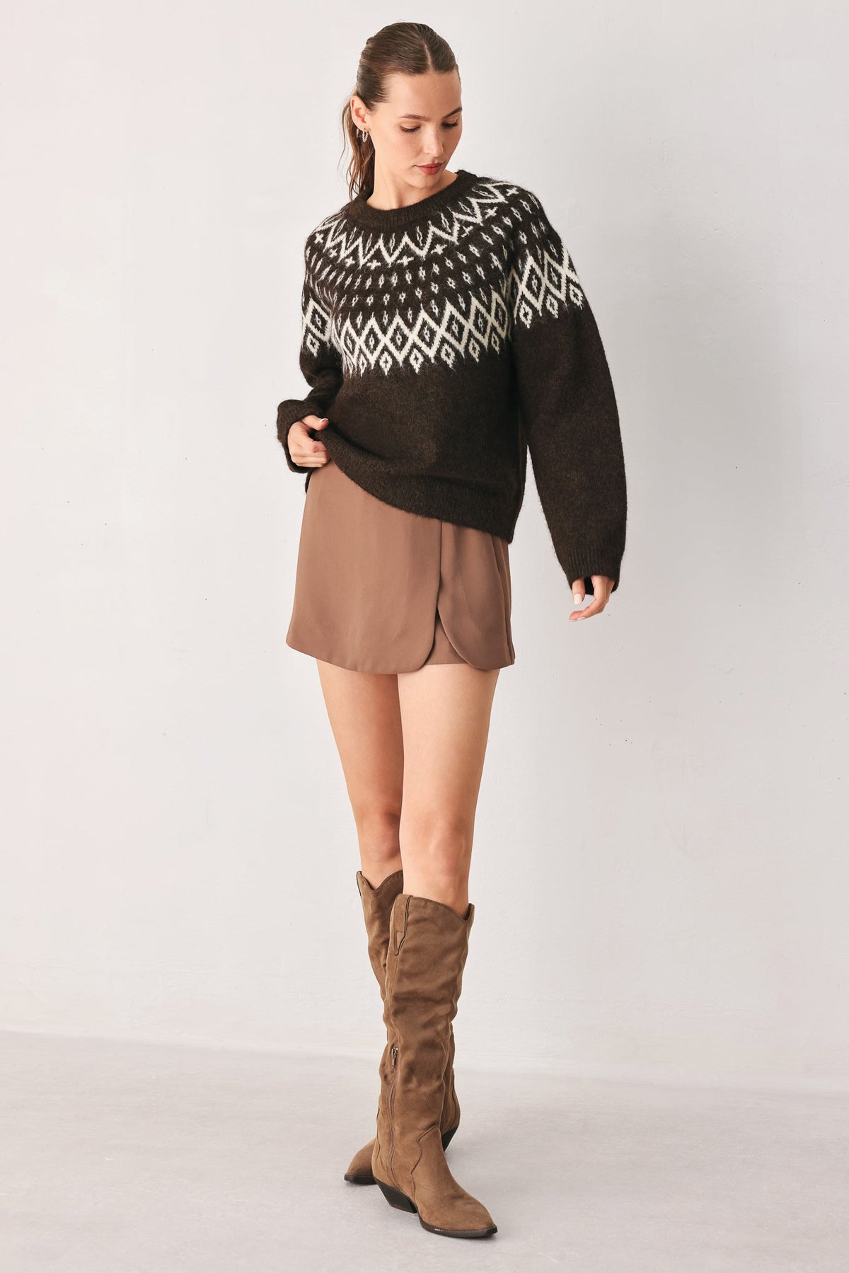 A woman modeling a dark brown knit sweater with a white geometric pattern across the chest and shoulders, paired with a brown mini skirt and brown over-the-knee boots, against a light gray studio background.