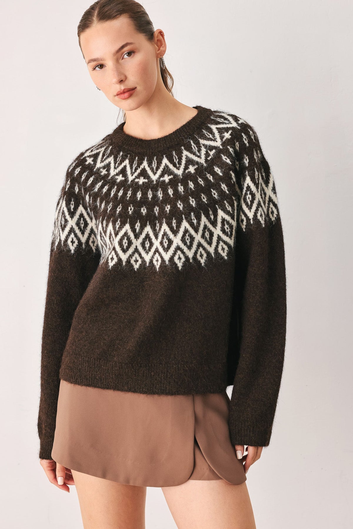 A woman wearing a dark brown knit sweater with a white geometric Fair Isle pattern across the chest and shoulders, paired with a brown skirt, against a light gray studio background.