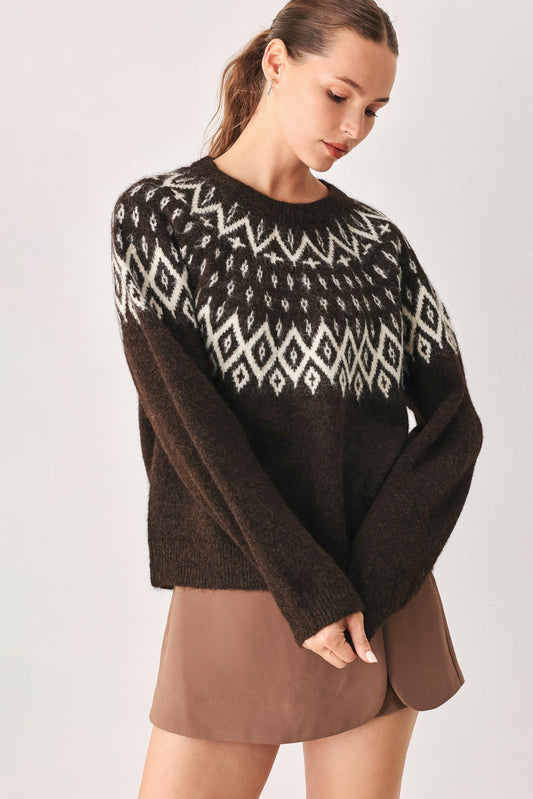 A woman wearing a dark brown knit sweater with a white geometric Fair Isle pattern across the chest and shoulders, paired with a brown mini skirt, against a light gray studio background.