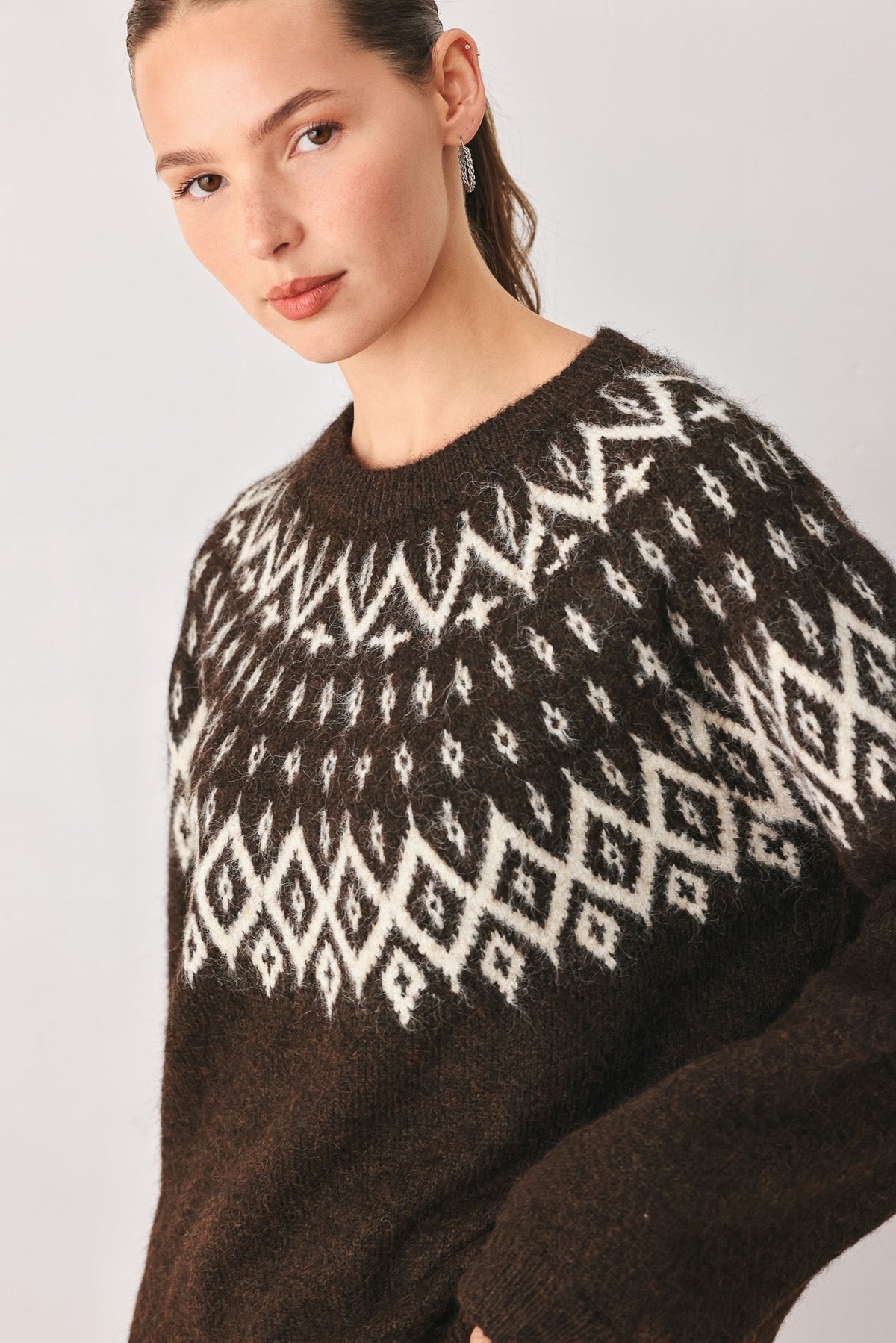A close-up of a woman wearing a dark brown fuzzy knit sweater with a cream-colored geometric Fair Isle pattern across the chest and shoulders, set against a plain light gray background.