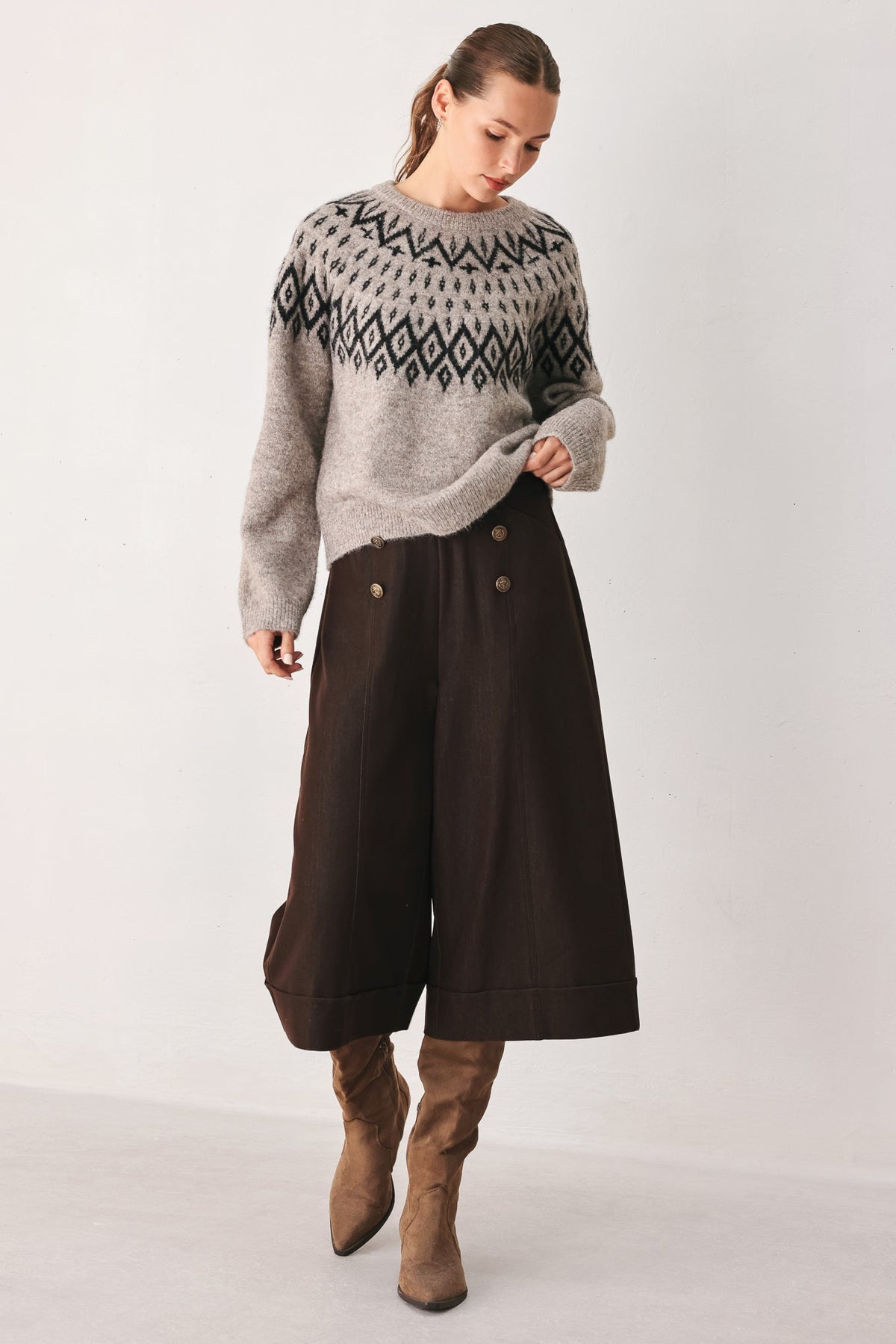 A woman modeling a light gray knit sweater with a black geometric pattern across the chest and shoulders, paired with dark brown wide-leg cropped pants and brown ankle boots, against a plain light gray studio background.