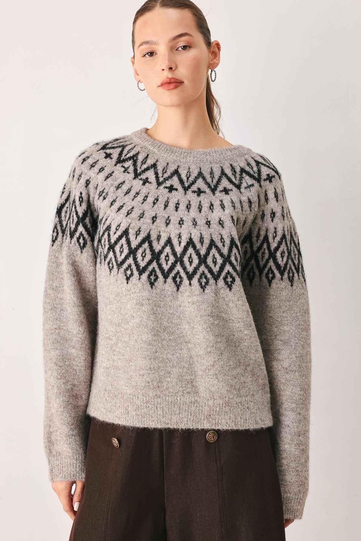 A woman wearing a light gray knit sweater with a black geometric Fair Isle pattern across the chest and shoulders, paired with dark brown pants, against a plain light gray studio background.