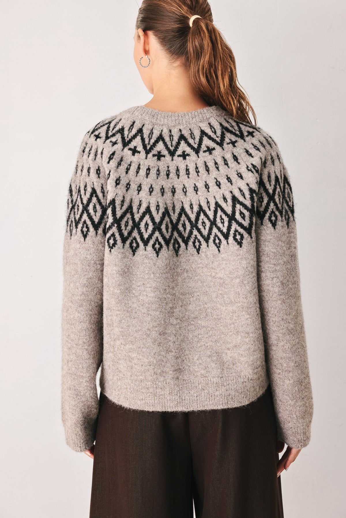 A woman viewed from the back wearing a light gray knit sweater with a black geometric Fair Isle pattern across the shoulders and upper back, paired with dark brown pants, against a plain light gray studio background.