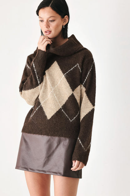 A woman wearing a brown and beige argyle knit sweater with a high folded collar, paired with a dark brown faux-leather mini skirt, against a plain white studio background.