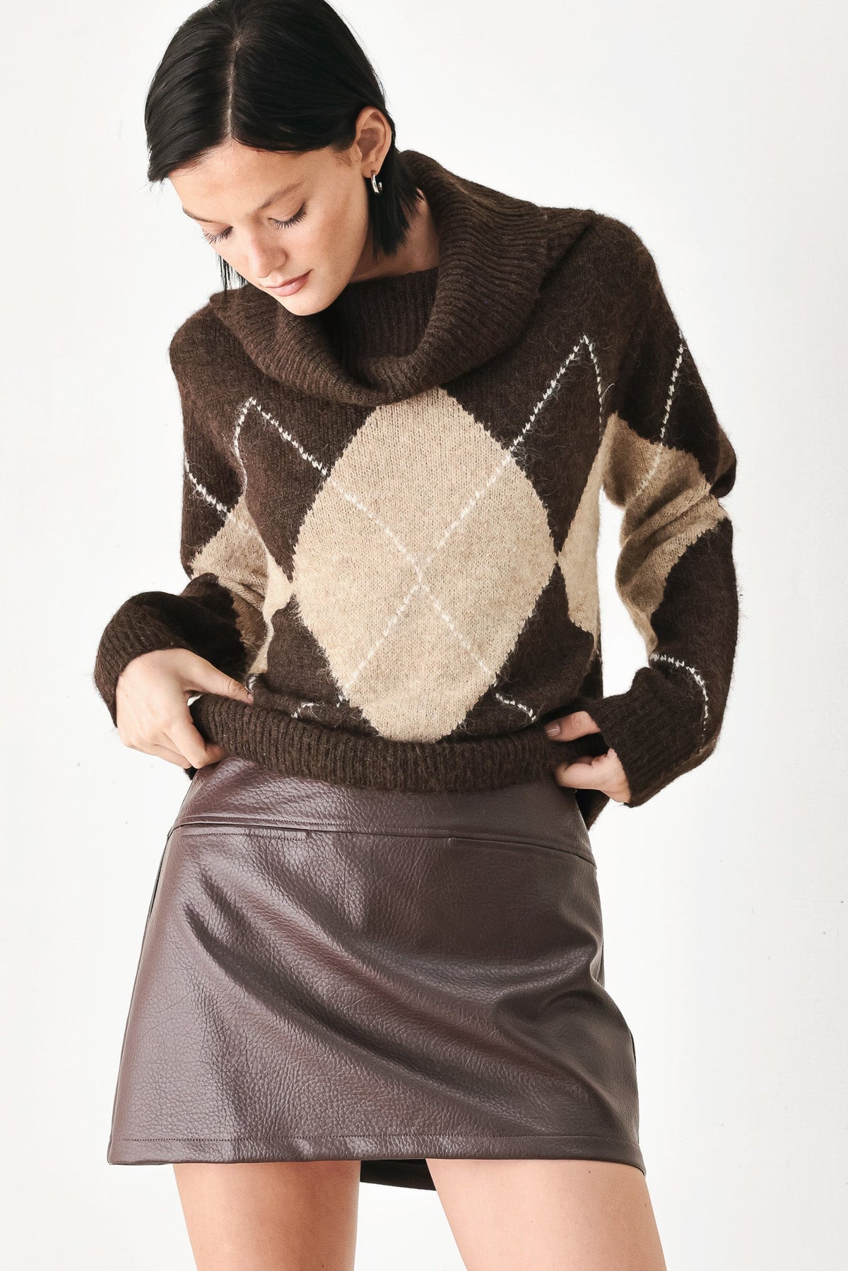 A woman wearing a brown and beige argyle knit sweater with a high folded collar, paired with a dark brown faux-leather mini skirt, against a plain white studio background.