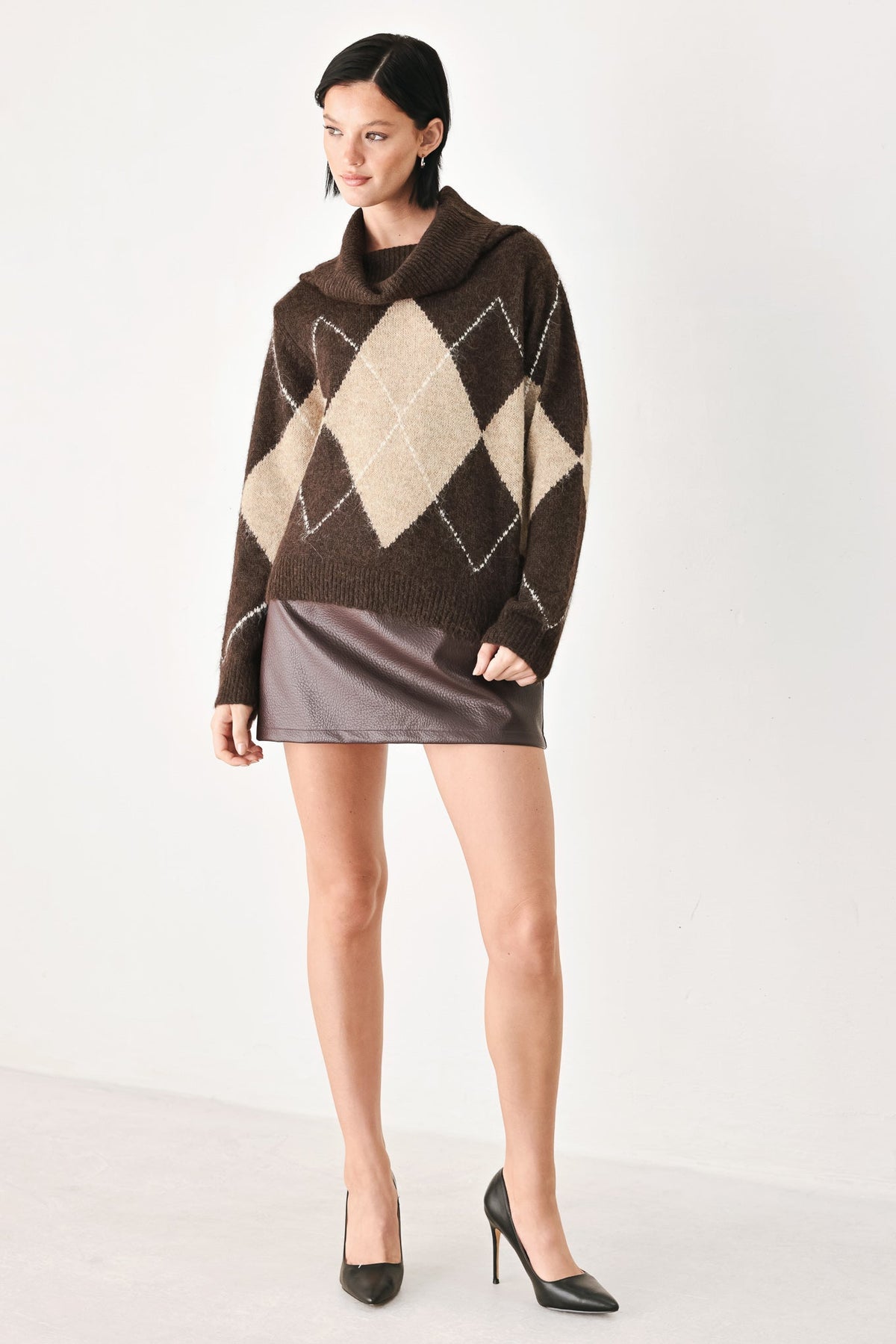 A model wearing a brown and beige argyle knit sweater with a cowl neck, paired with a brown mini skirt and black high heels, set against a plain white studio background.