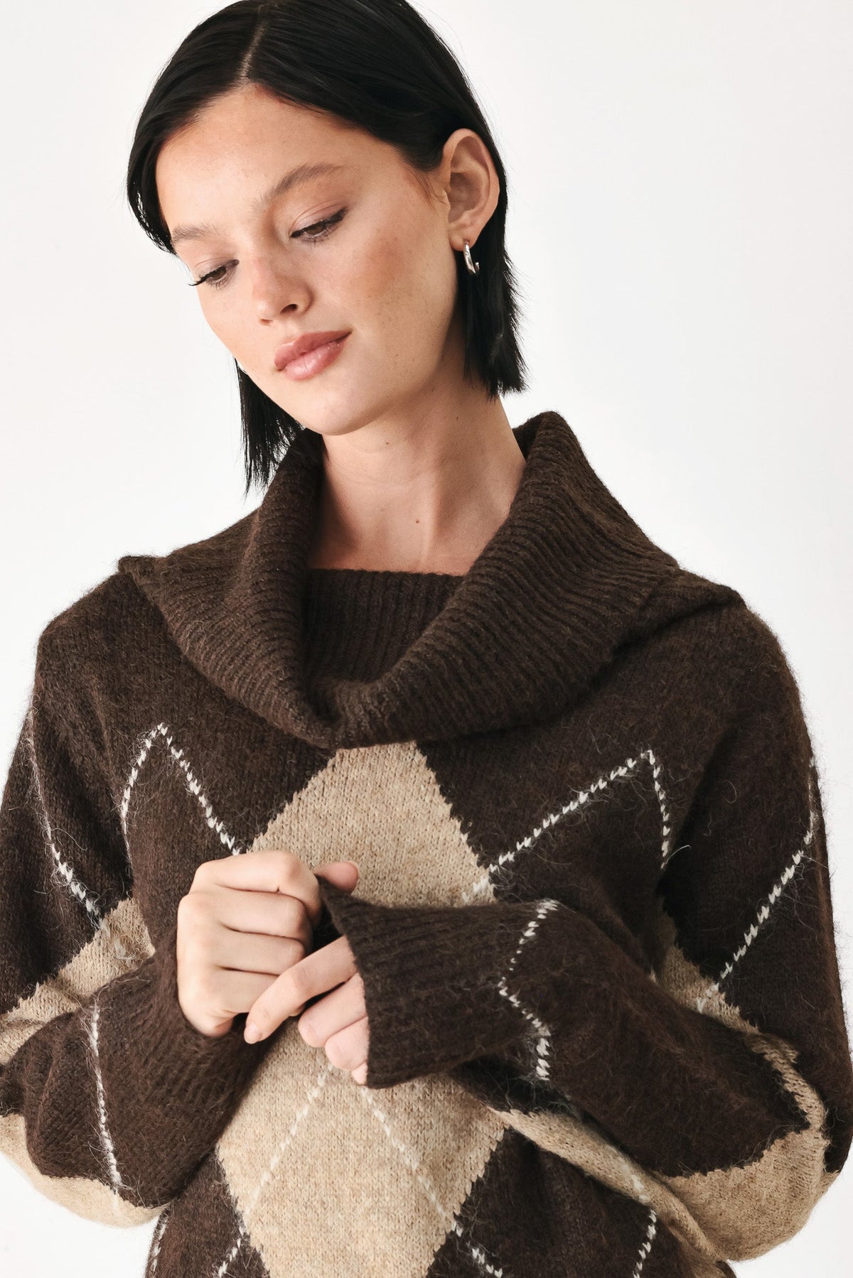 A woman wearing a brown and beige argyle knit sweater with a high ribbed cowl neck, shown from the chest up against a plain light background.