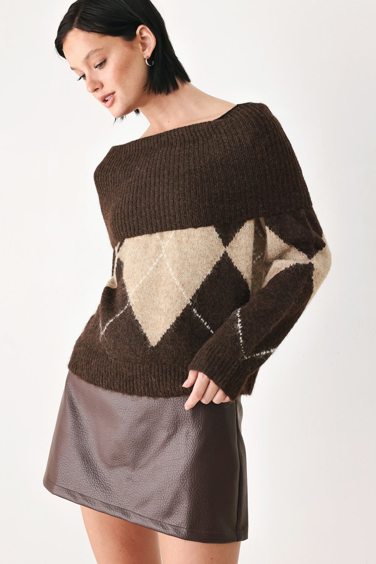 A woman wearing a dark brown off-the-shoulder knit sweater with a beige and brown argyle pattern on the front, paired with a dark brown faux-leather mini skirt, against a plain white studio background.