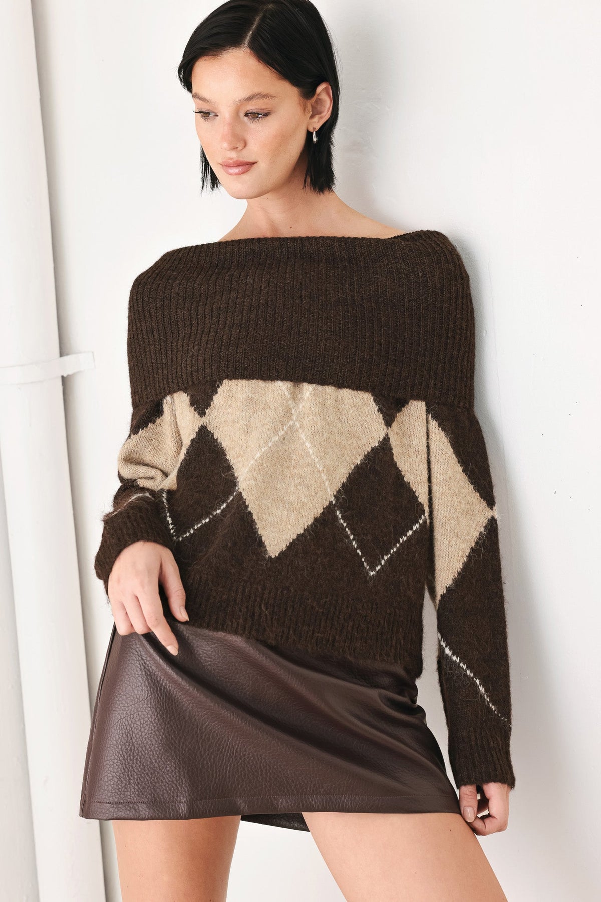 A woman wearing a dark brown off-the-shoulder knit sweater with a beige and brown argyle pattern on the body and sleeves, paired with a dark brown faux-leather mini skirt, standing against a white wall.