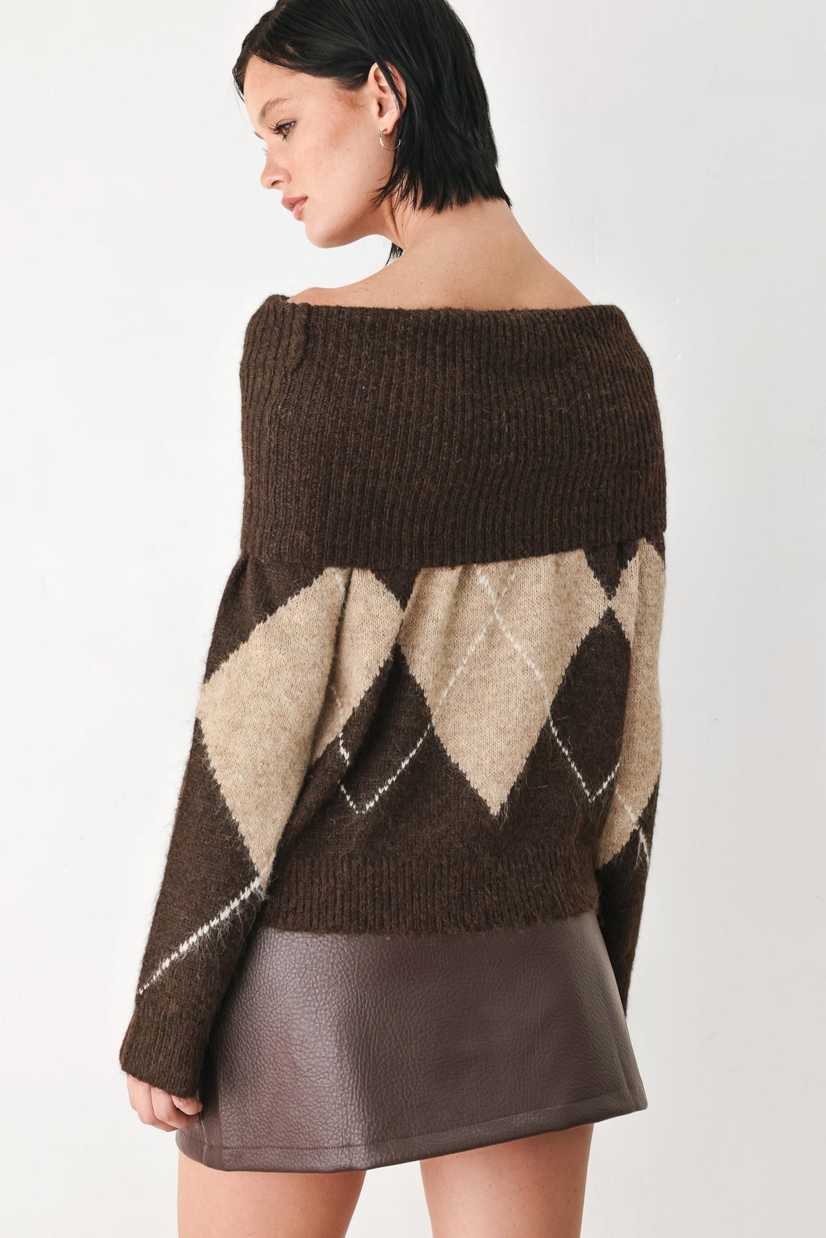 A woman wearing a brown off-the-shoulder knit sweater with a beige and brown argyle pattern on the sleeves, paired with a brown faux-leather mini skirt, shown from a side-back angle against a light gray studio background.